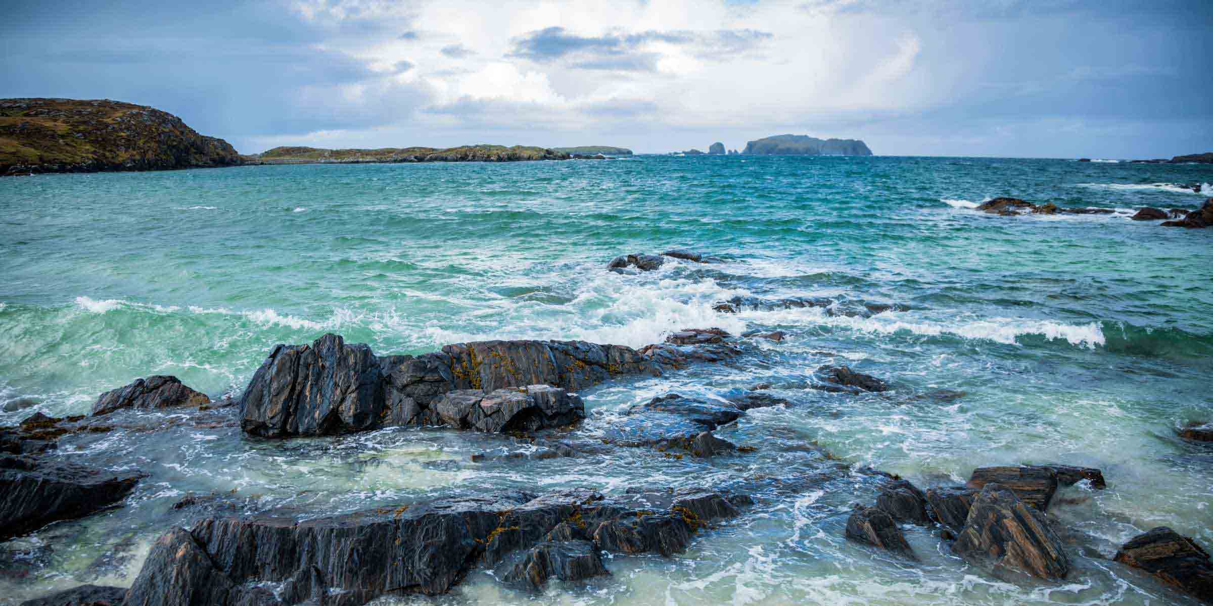 Rugged coastal landscape of the Outer Hebrides with turquoise waves crashing against rocky shores and distant islands under a blue sky with whispy clouds.