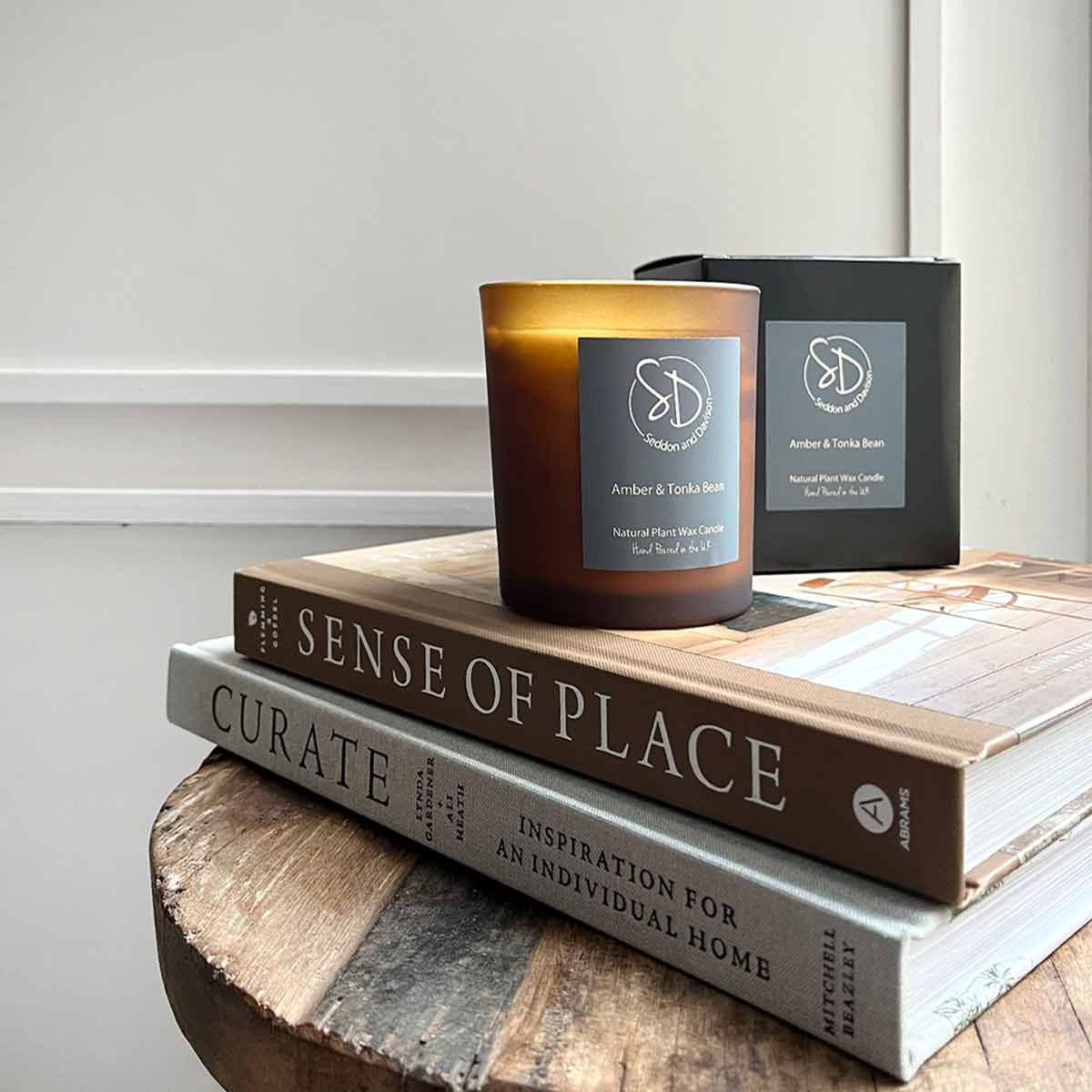 A 30cl Black Pomegranate Scented Candle in a frosted amber glass jar, placed on stacked interior design books, with its matching black packaging in the background.